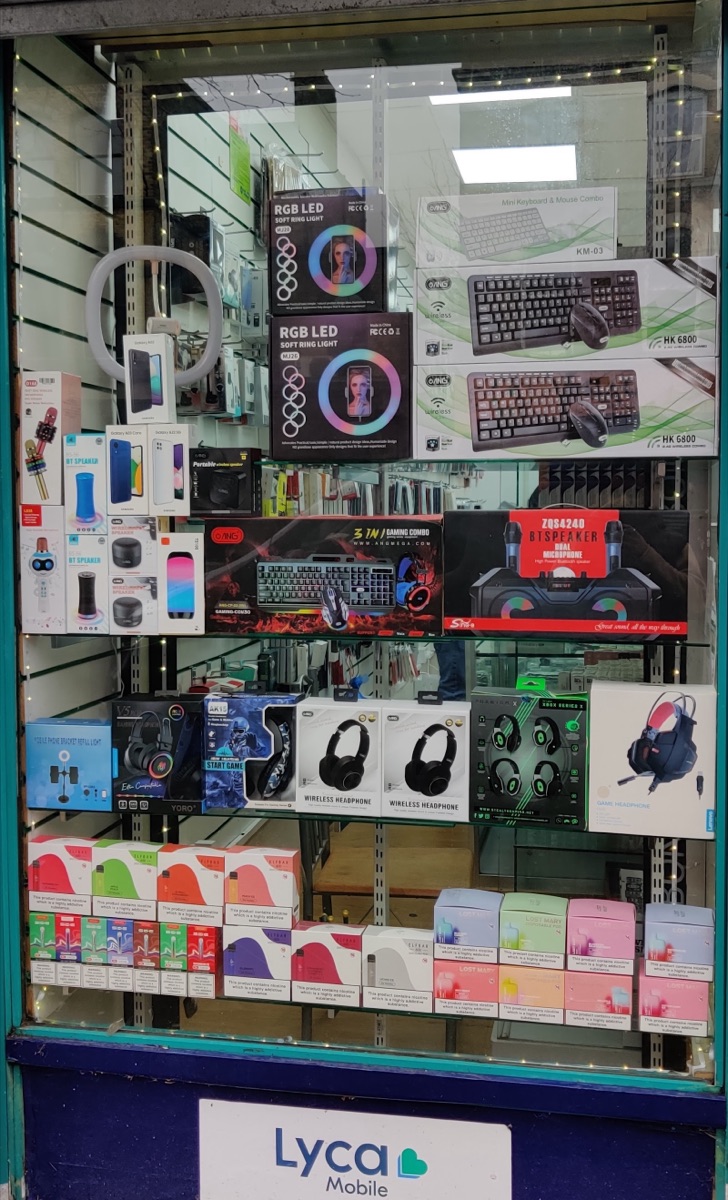 Window display at Sid Mobiles packed with speakers, headphones, ring lights, keyboards and vapes.